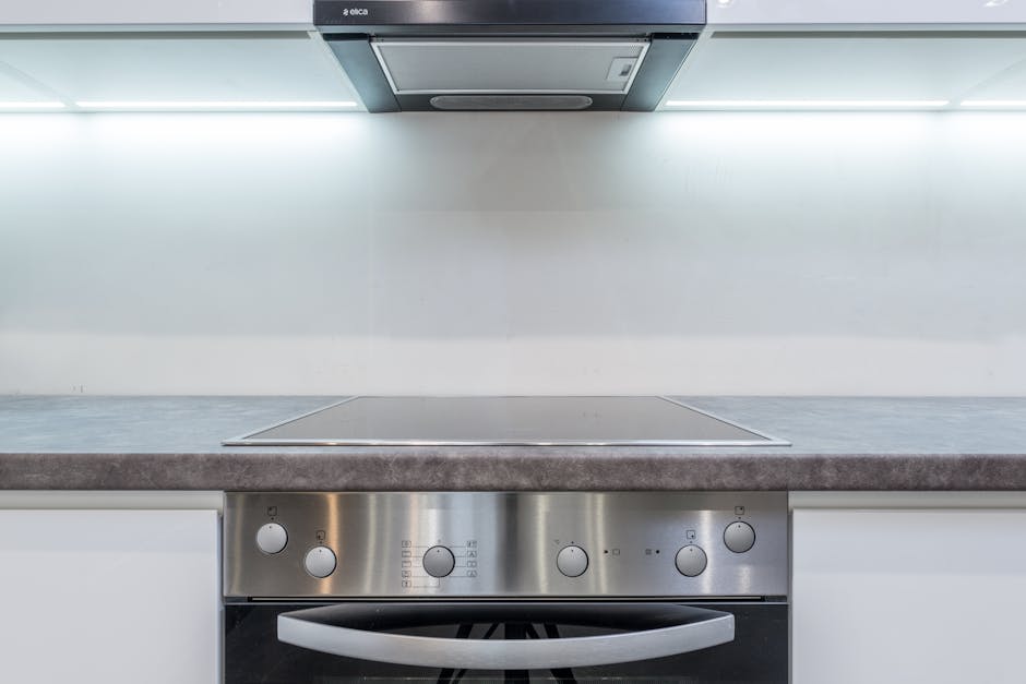 A commercial kitchen sink area with stainless steel surfaces, featuring a stainless steel sink with a flexible coiled hose and a cleaning cloth hanging on it. The tiled wall behind is light blue with 