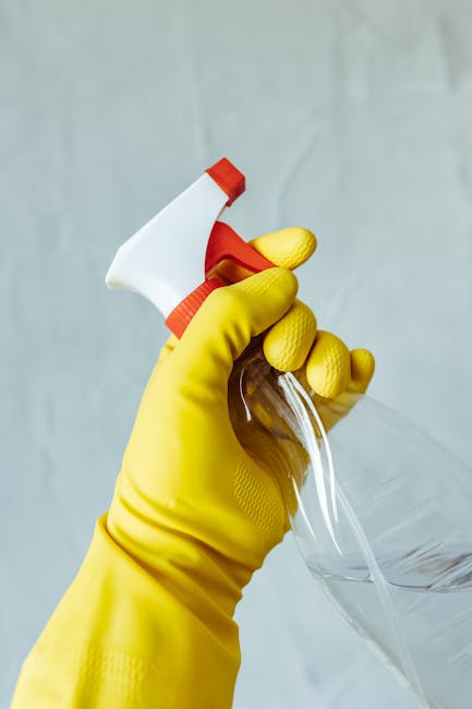 A person wearing bright yellow rubber gloves is cleaning a white surface with a blue sponge held in one hand, while the other hand, also gloved, holds a spray bottle of clear cleaning solution. The su