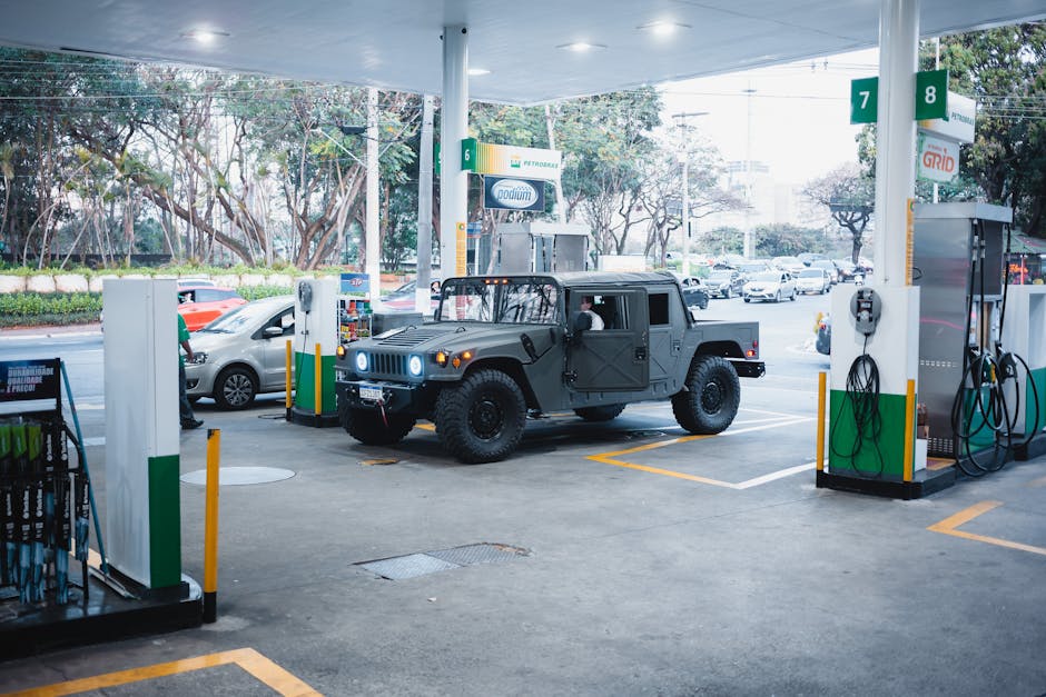 A grey military-style vehicle parked at a petrol station with green and white fuel pumps, surrounded by other cars and trees in the background. The station has a white canopy overhead, with visible fu